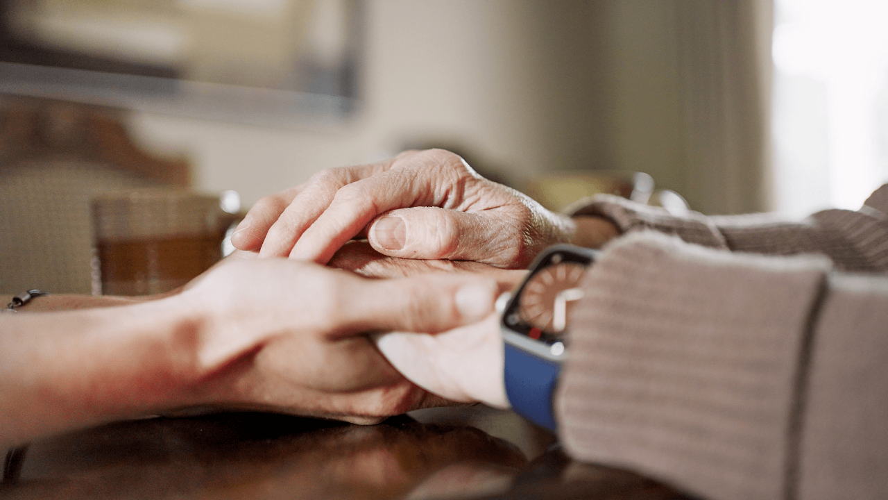 Close-up of a young woman holding her mature mother's hands in support at a table during a visit